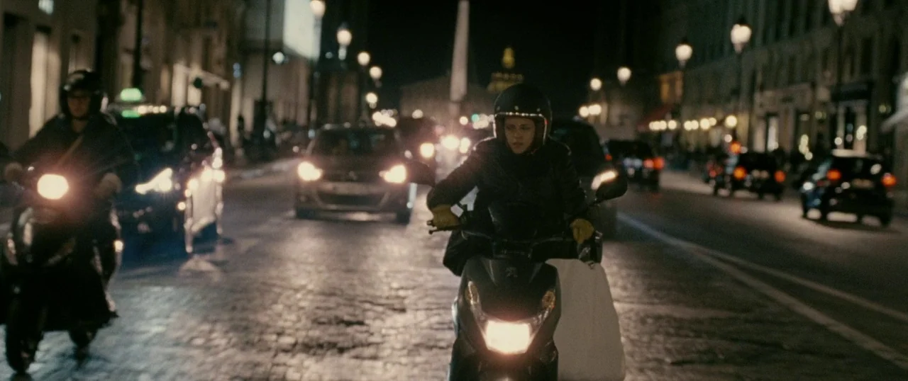 Personal Shopper (2016) movie still: medium — A woman drives a scooter through a cobblestone city street at night. Numerous cars and another scooter…