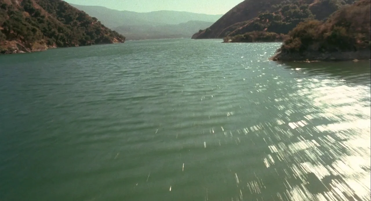 Koyaanasquatsi (1982) movie still: establishing — A reservoir surrounded by arid, brown, shrub-covered hills sits under a clear sky. The surface of…