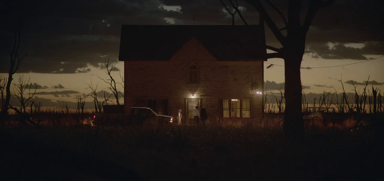 Foe (2023) movie still: wide — A weathered farmhouse sits in a vast, dark field under a dim twilight sky. Three people stand gathered at the…