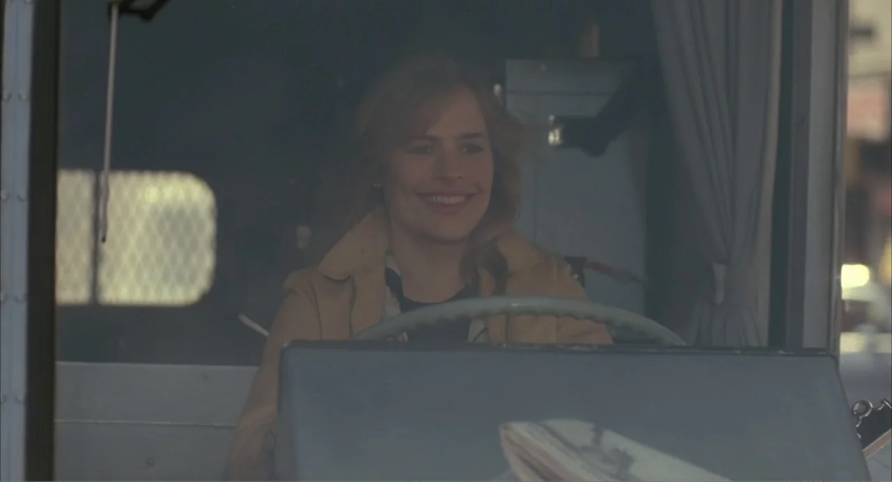 F/X (1986) movie still: medium — A woman sits in the driver's seat of a truck with fabric curtains visible. The driver smiles while holding the…