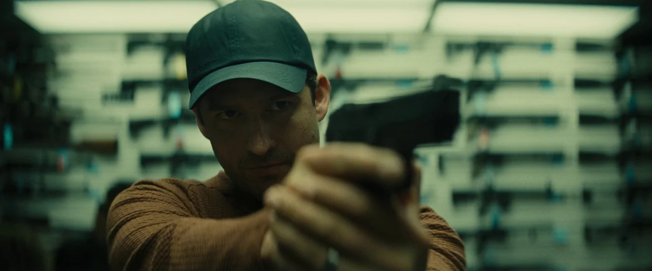 Knock at the Cabin (2023) movie still: closeup — Rows of firearms are mounted on the wall of a dimly lit store interior. A man wearing a baseball…
