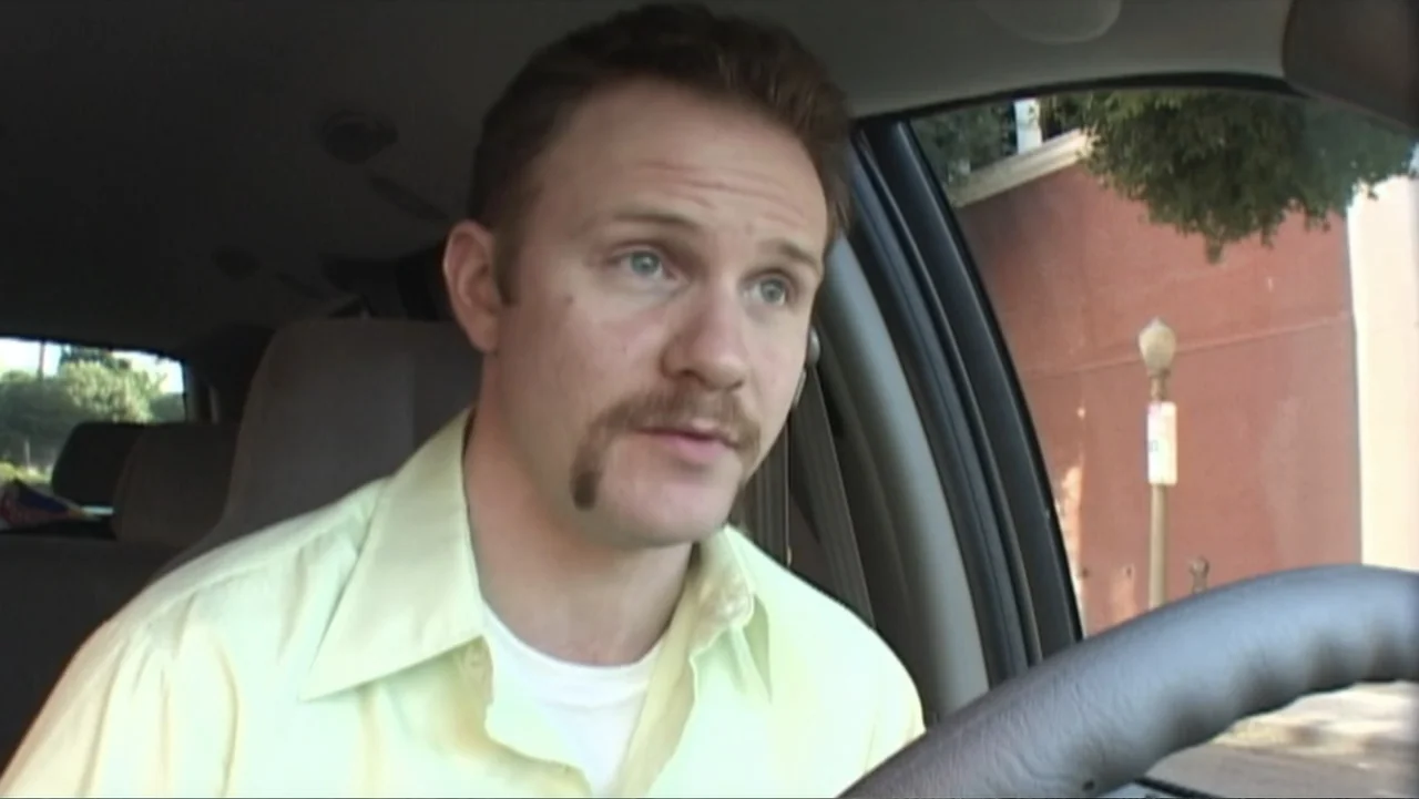 Super Size Me (2004) movie still: medium — A man sits in the driver seat of a car during the day. He is turned toward the passenger side while…