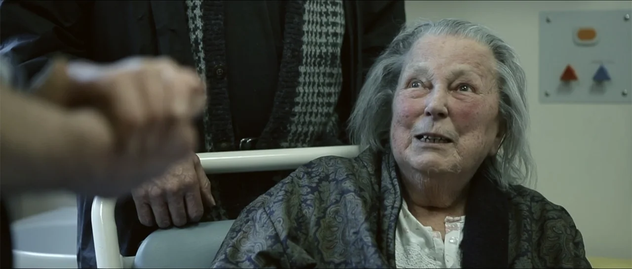 Garnet’s Gold (2014) movie still: medium — A hospital room with a white wall panel featuring colored buttons and switches. An elderly woman sits in…