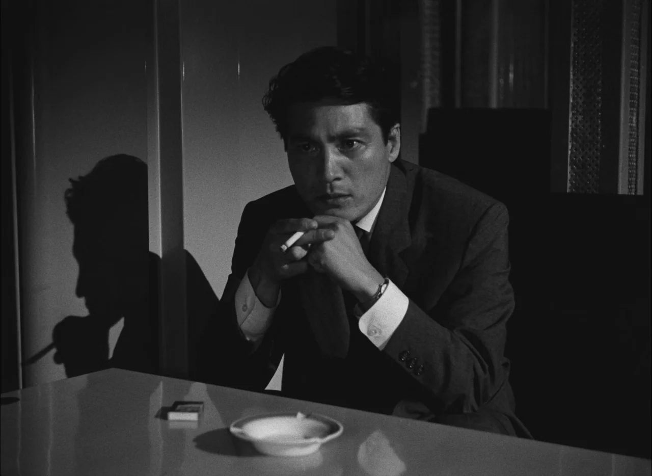 Hiroshima Mon Amour (1959) movie still: medium — A dark, spartan room features a single desk with a clean white surface. A man sits at the desk with…