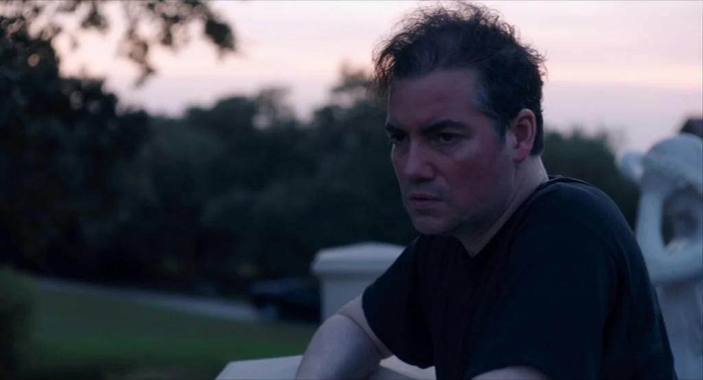 Results (2015) movie still: medium — A stone railing overlooks a blurred landscape during the twilight hours of the day. A middle-aged man leans on…