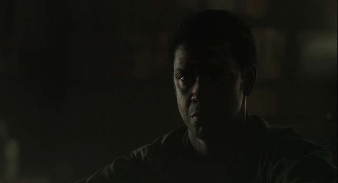 American Gangster (2007) movie still: closeup — A man sits in a dark, indistinct interior setting. He gazes forward while illuminated by a dim,…