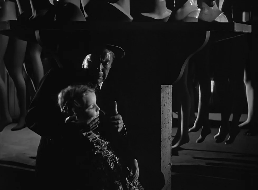 M (1951) (1951) movie still: medium — A dark, cluttered mannequin storage room filled with hanging legs and mannequin heads. A man stands behind a…