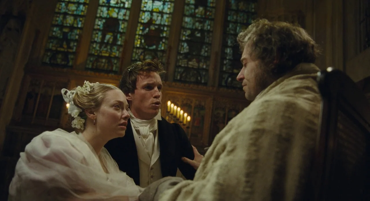 Les Misérables (2012) movie still: medium — A dimly lit church interior features ornate stained glass windows and wooden paneling. A young man and…