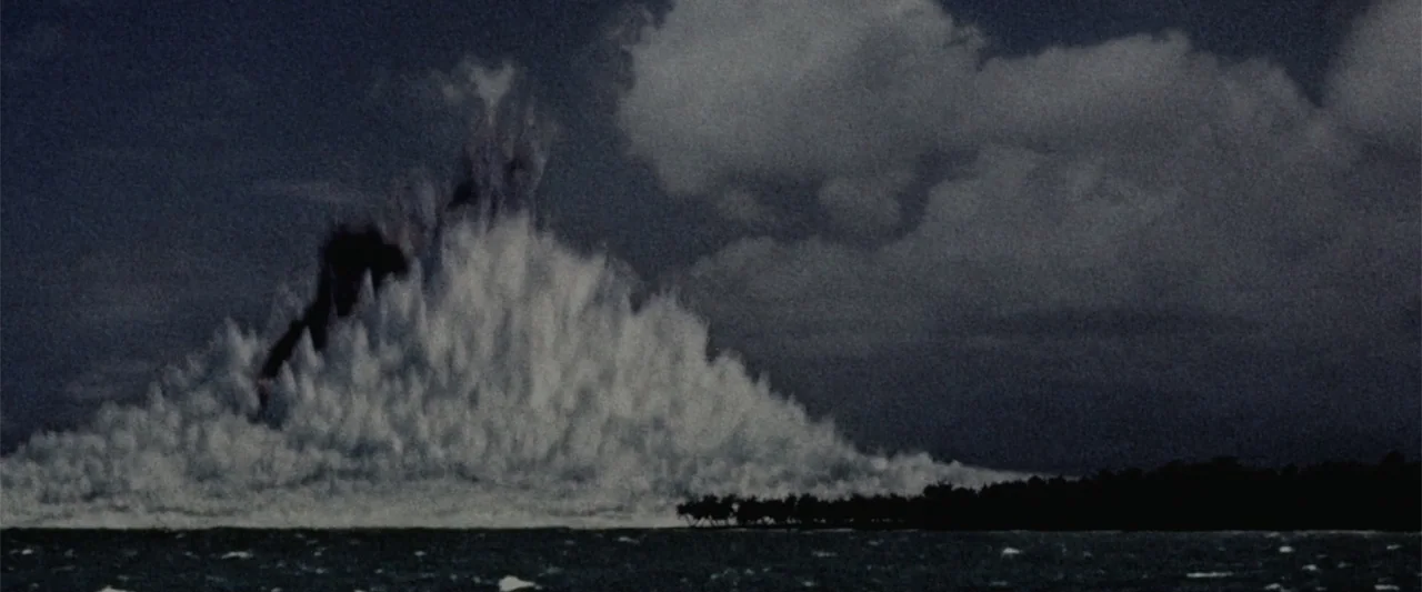 Godzilla (2014) movie still: wide — An explosive white water geyser rises against a dark, overcast sky above the ocean. A thin sliver of a tropical…
