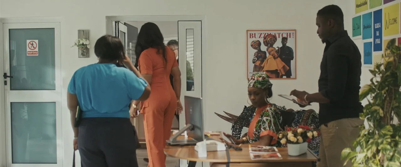 Casa De Novia (2024) movie still: medium — A modern reception area features white walls, a wooden desk, and a framed magazine poster. Several people…