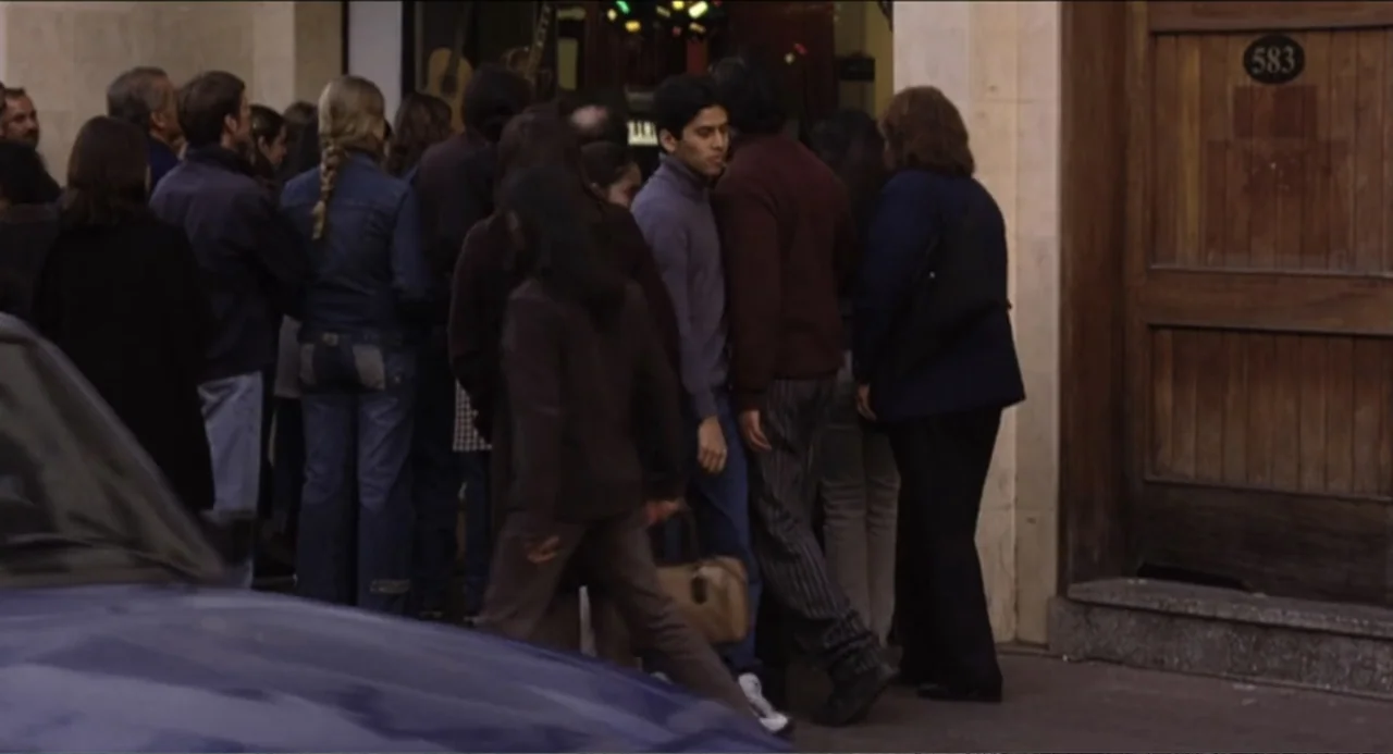 The Holy Girl (2004) movie still: medium — A group of people gathers on a sidewalk in front of a wooden door. Several individuals in casual winter…