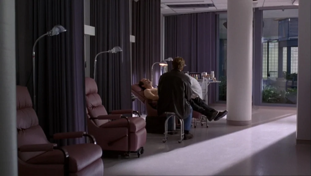 Crash (1996) movie still: wide — A clinical medical bay features rows of purple vinyl recliners separated by heavy curtains. Two men occupy the…