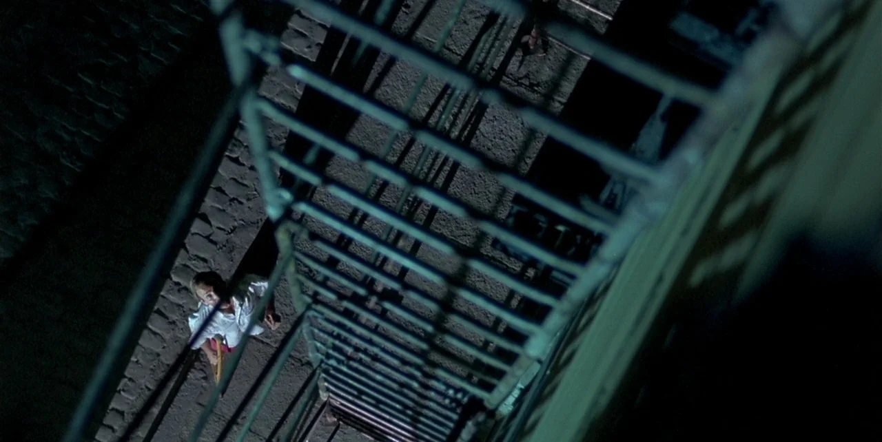 Law of Desire (1987) movie still: wide — A top-down view looks down a metal fire escape toward a dark urban alleyway. A man stands on the ground…