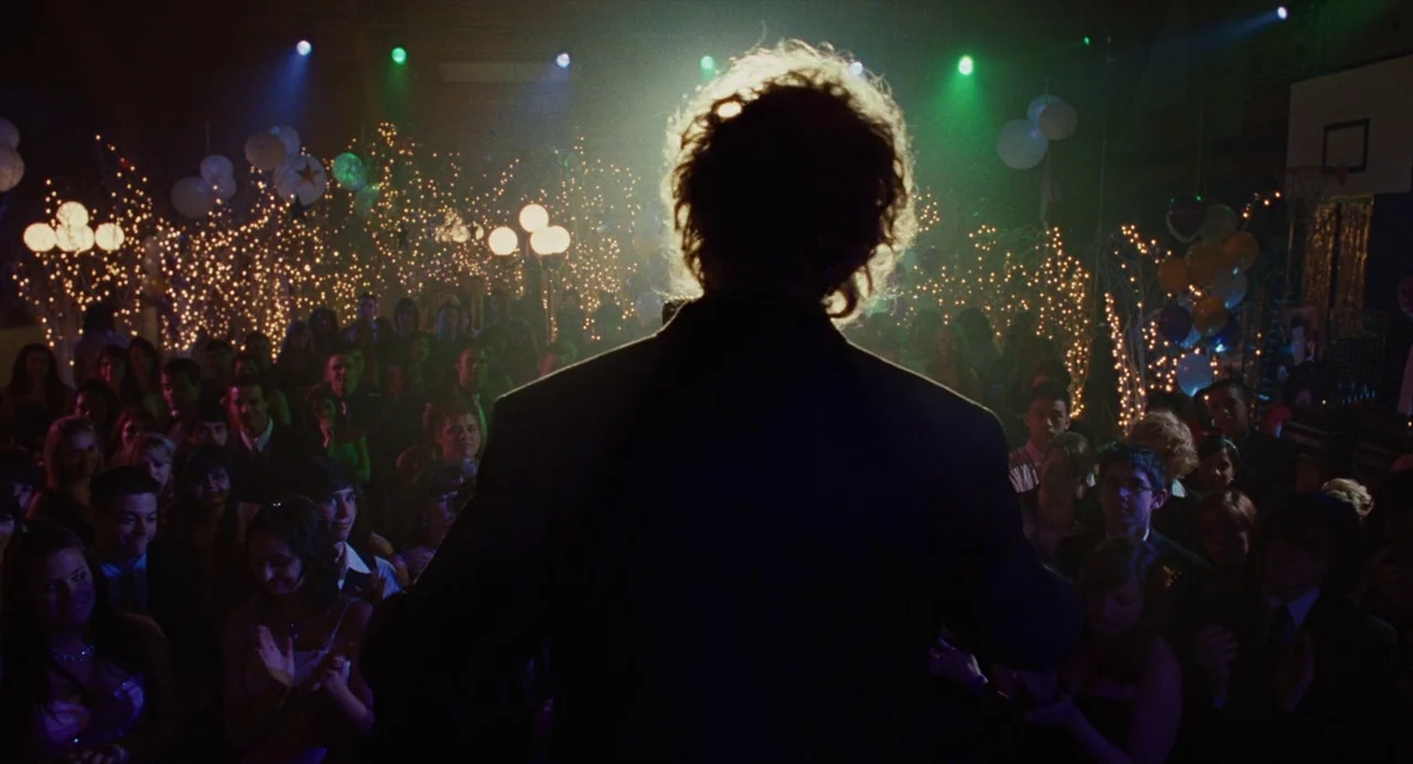 Jennifer’s Body (2009) movie still: medium — A dark, crowded gymnasium filled with fairy lights and balloons. A young man in a suit stands in the…
