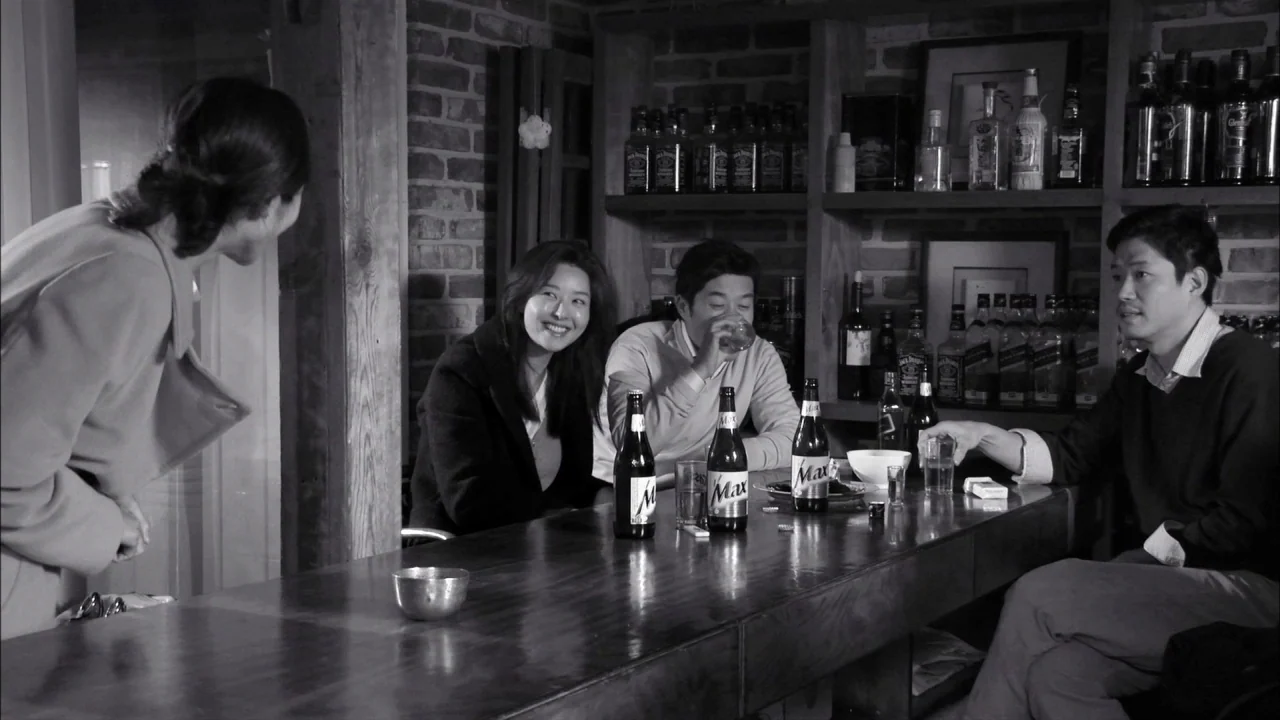 The Day He Arrives (2018) movie still: medium — A dimly lit counter area features brick walls and shelves stocked with alcohol bottles. Three people…
