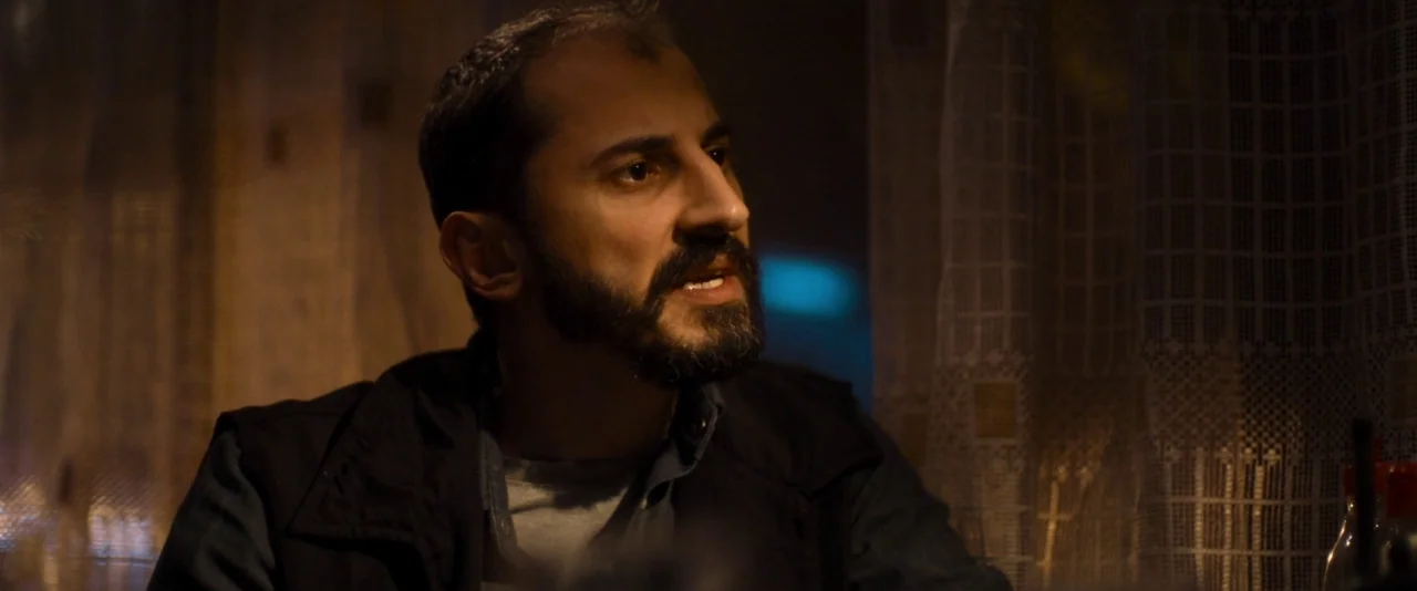 Baskin (2015) movie still: medium — A dimly lit room features patterned lace curtains hanging in the background. A man with a beard sits and speaks…