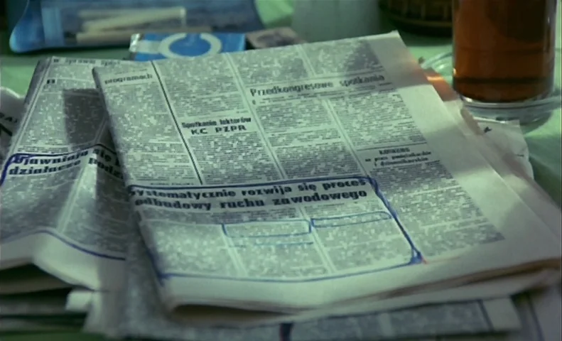 No End (1985) movie still: closeup — Polish newspapers with blue ink markings lie on a light-colored table surface. A glass of amber-colored liquid…