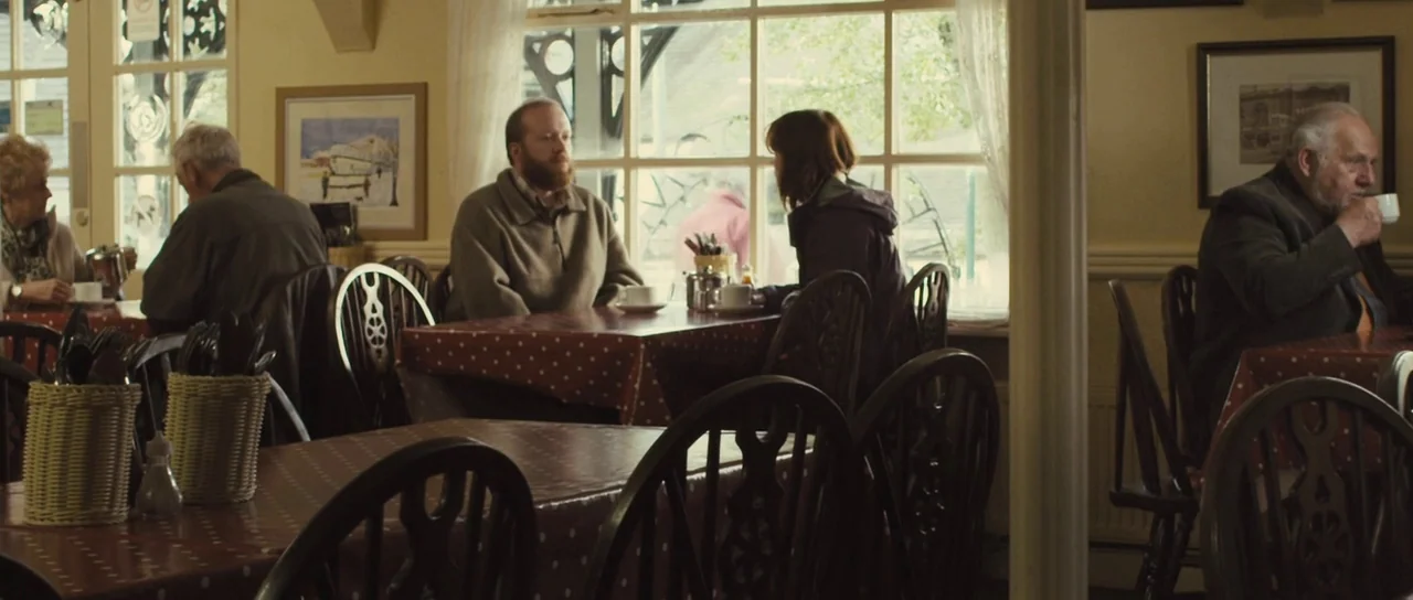 Sightseers (2012) movie still: wide — A quiet cafe interior features wooden chairs and tables covered with red polka-dot tablecloths. Several people…