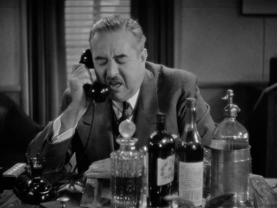 It Happened One Night (1934) movie still: medium — A man in a suit sits at a desk containing several glass liquor bottles. He talks on a black…