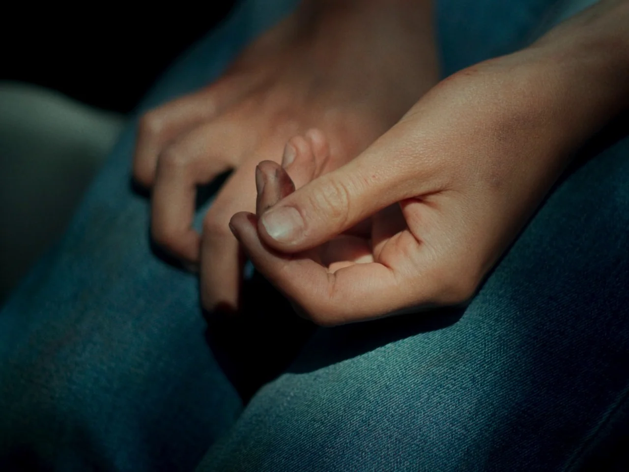 In A Violent Nature (2024) movie still: closeup — The hands rest on a pair of blue denim jeans. The fingers have visible dark smudge marks on them.
