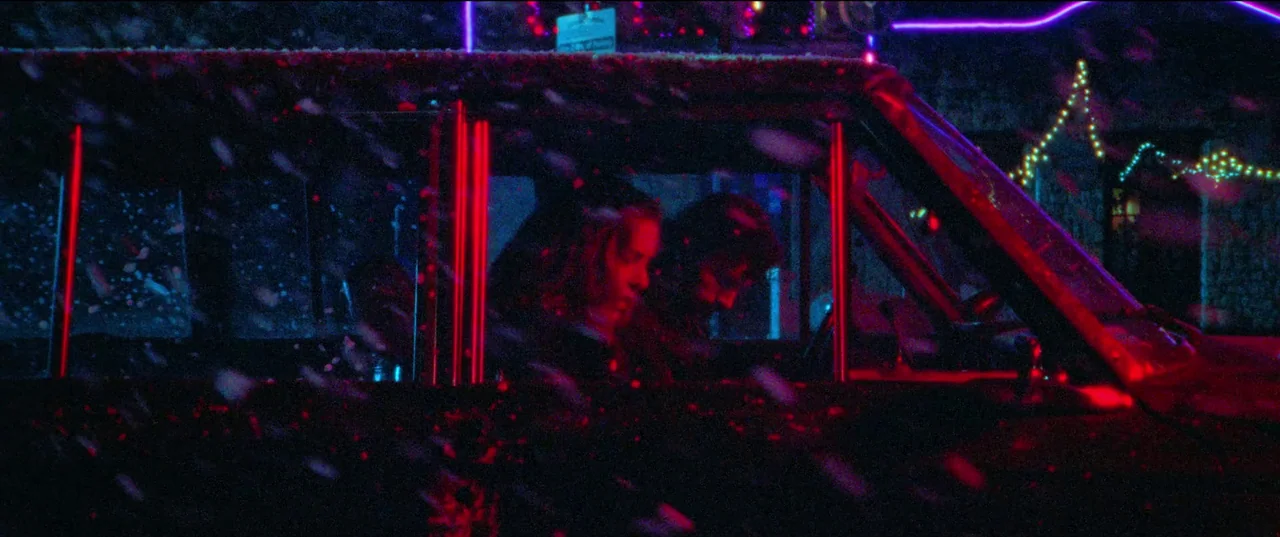 Christmas Bloody Christmas (2022) movie still: medium — A car parked in a snow-covered area at night illuminated by bright red neon lights. Two…