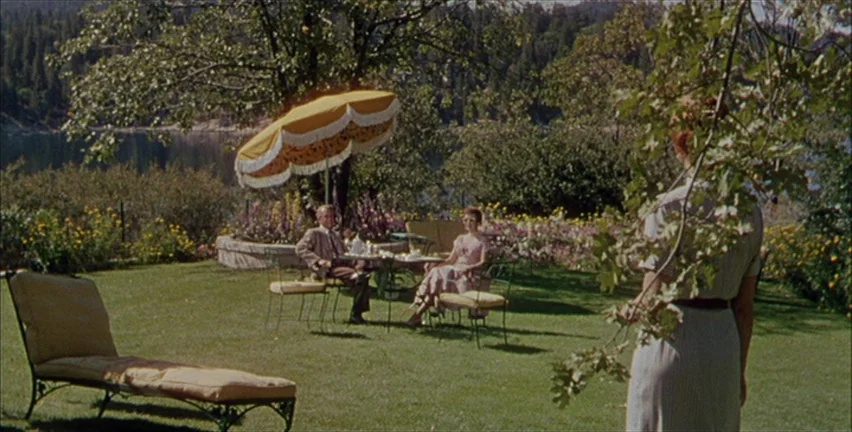 Magnificent Obsession (1954) movie still: wide — A manicured lakeside lawn features a dining table with a yellow fringe umbrella. A woman stands in…