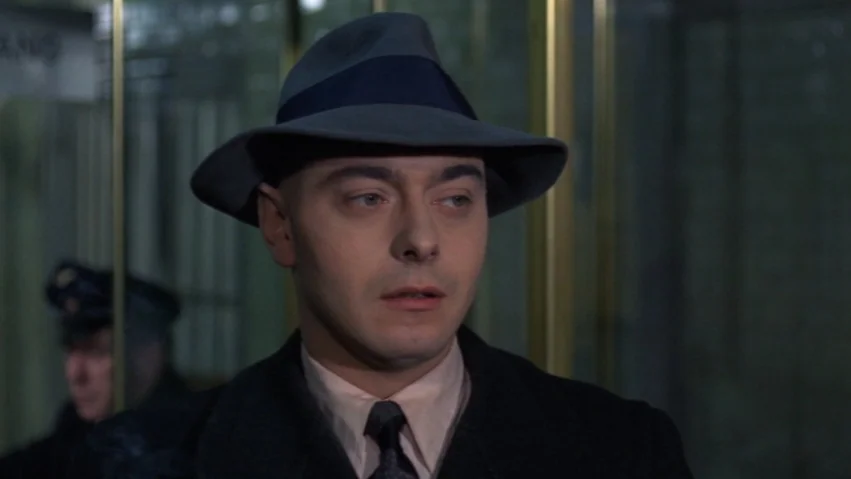 Tenderness of the Wolves (1973) movie still: closeup — A man stands inside a building structure with gold-framed glass panels behind him. He wears a…