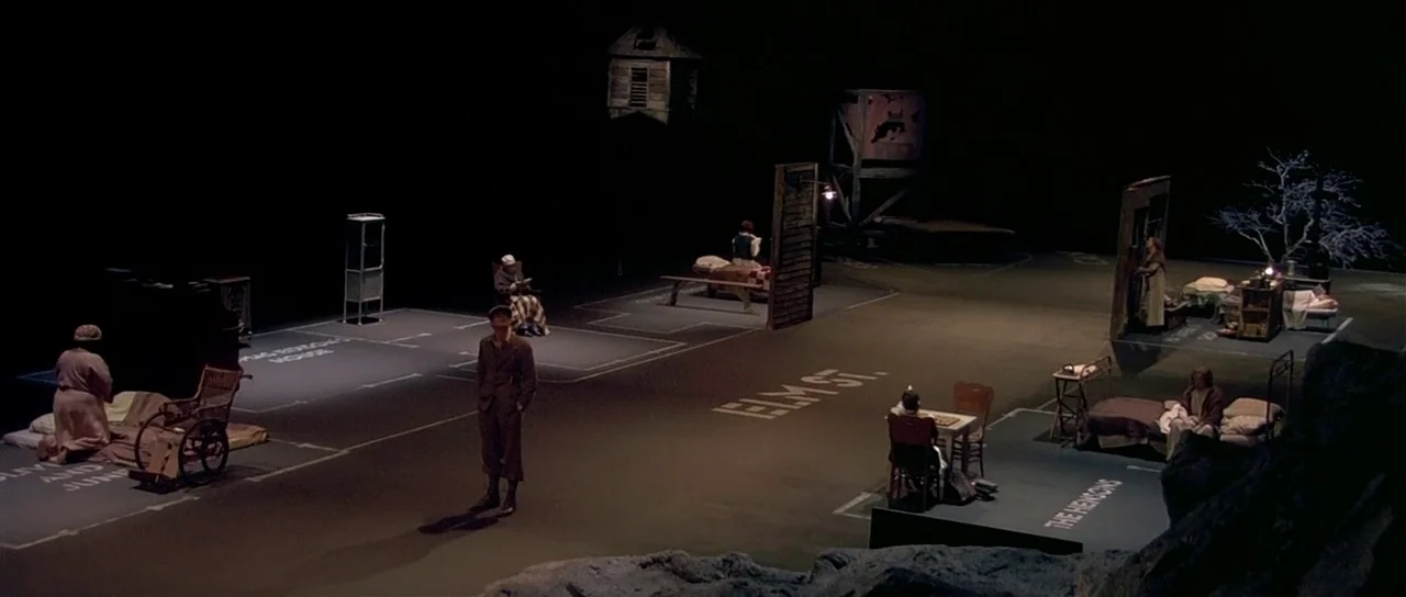 Dogville (2003) movie still: wide — A dark theatrical stage features outlined room floor plans drawn directly onto the ground. Multiple actors…