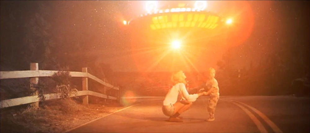 Close Encounters Of The Third Kind (1977) movie still: wide — A rural asphalt road curves past a wooden post-and-rail fence at night. A large,…