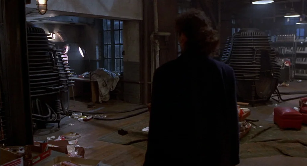 The Fly (1986) movie still: medium — A cluttered industrial loft features two large, metallic heat-sink-like structures flanking the room. One man…