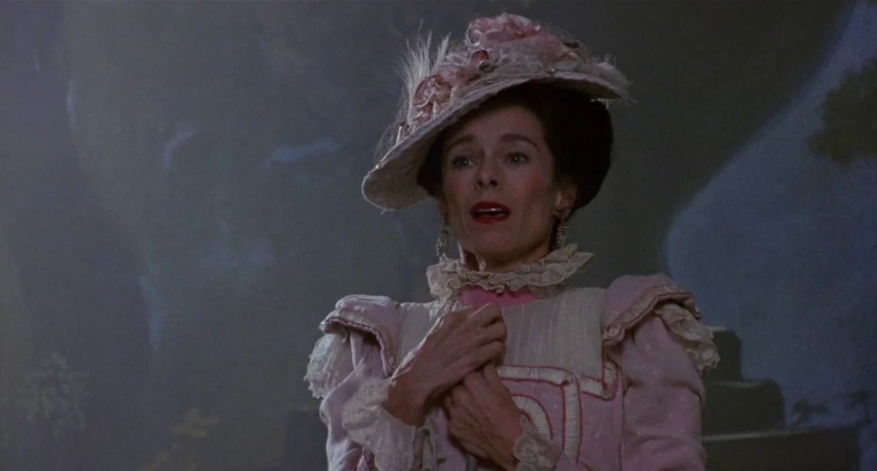 Chaplin (1992) movie still: medium — A woman stands in a dimly lit interior space with shadows cast behind her. She wears a formal pink…