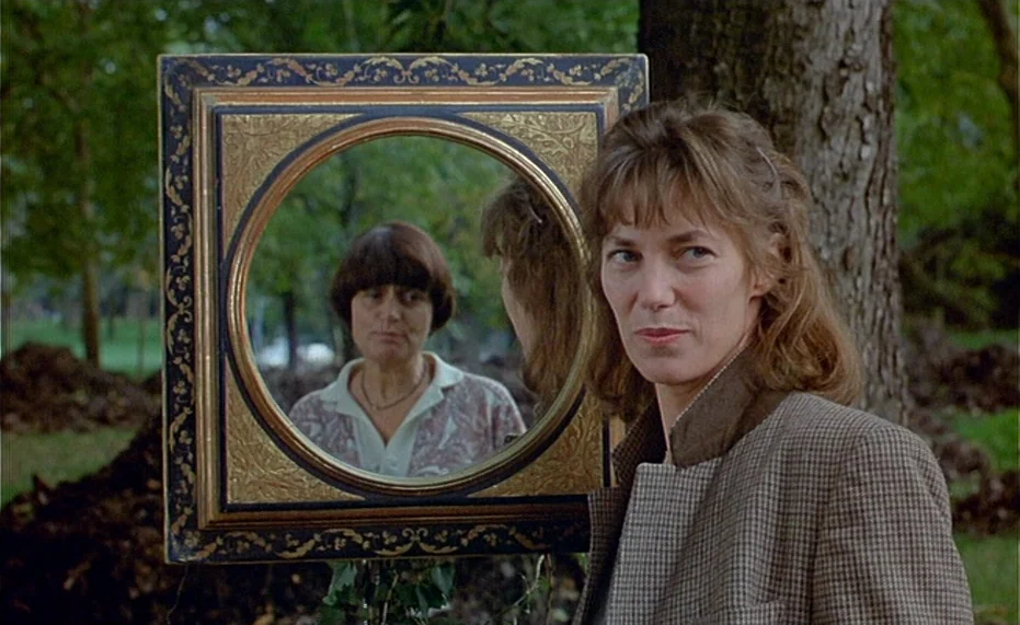 Jane B. par Agnès V. (1988) movie still: medium — A park setting with trees and dark soil visible behind a wooden-framed mirror. One woman stands in…