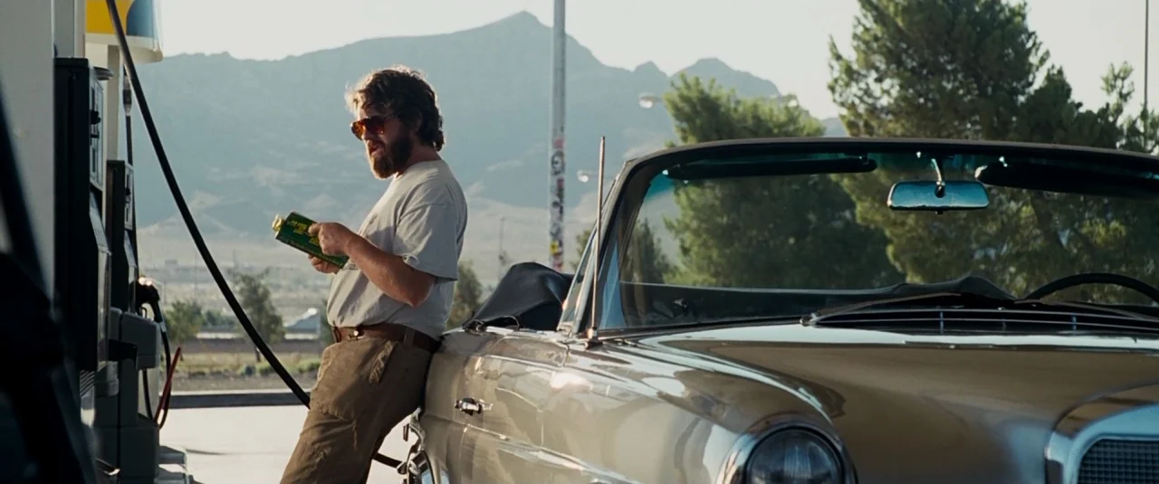 The Hangover (2009) movie still: medium — A man stands at a gas pump in a desert landscape during the day. He leans against a vintage convertible…