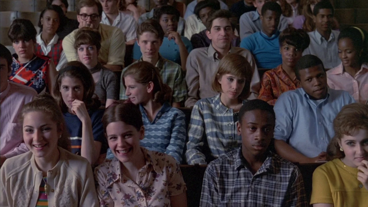 Baby It’s You (1983) movie still: medium — A crowded auditorium filled with young people in 1960s style clothing and hairstyles. The students sit in…