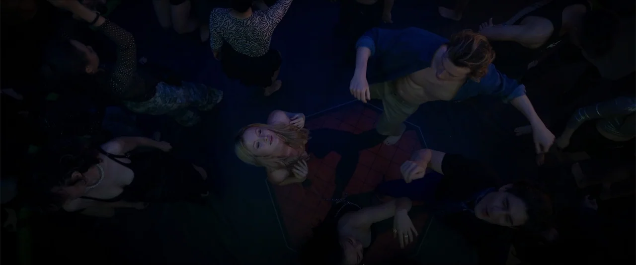 Love Me Love Me (2026) movie still: wide — A dark, crowded area features a lit geometric panel on the floor. Several individuals dance in the dim…