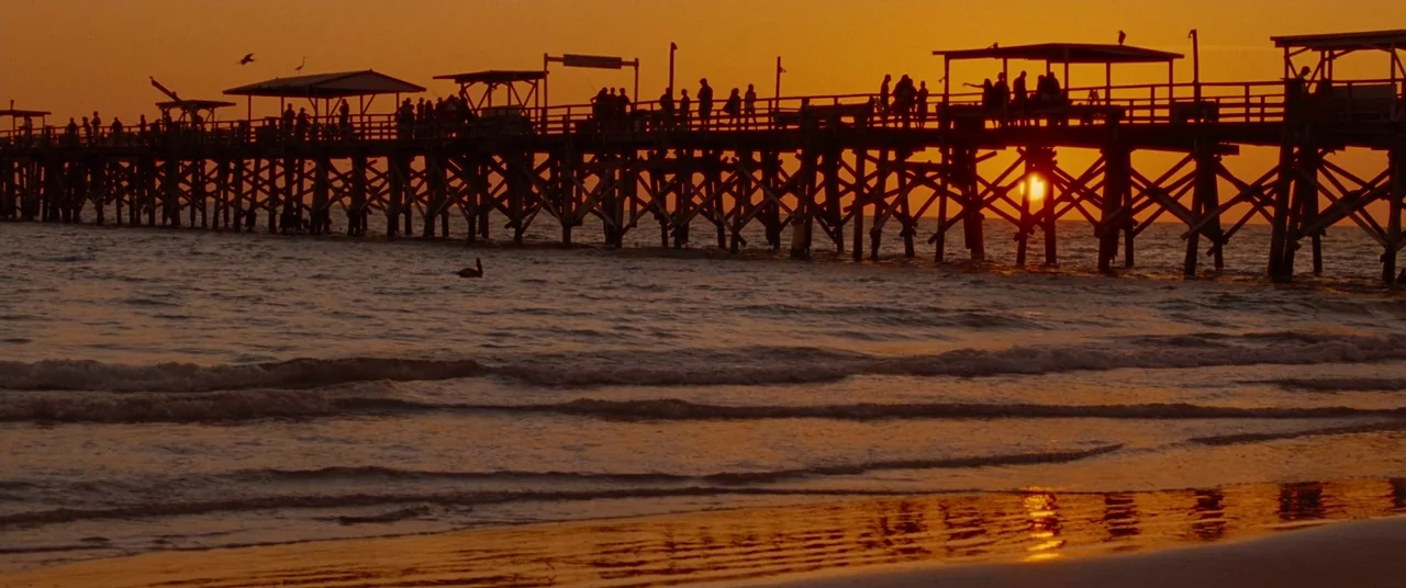 Spring Breakers (2012) movie still: wide — A long wooden pier extends across the frame over a wavy ocean at sunset. Silhouetted figures of many…