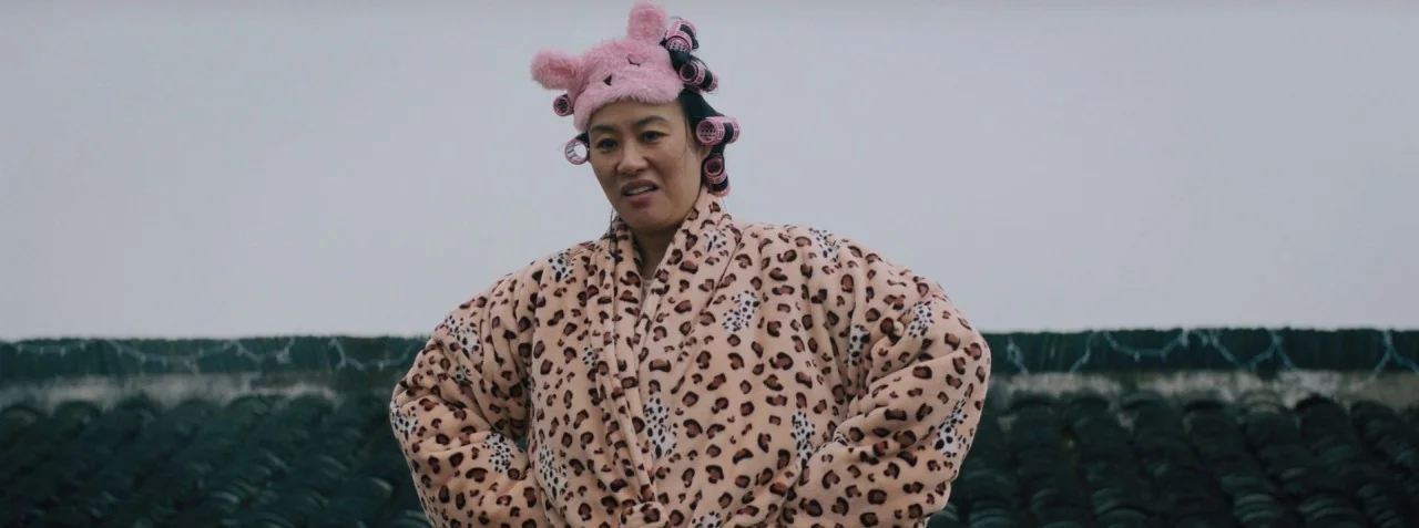 Dead Pigs (2018) movie still: medium — A woman stands in a large outdoor space filled with hundreds of stacked black tires. She wears a leopard…
