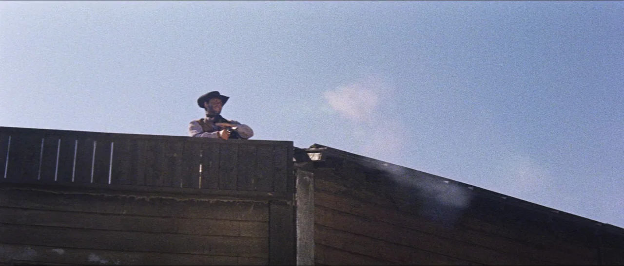 Texas, Adios (1966) movie still: wide — A man stands atop a wooden roof against a clear bright blue sky. He aims a handgun forward while smoke…