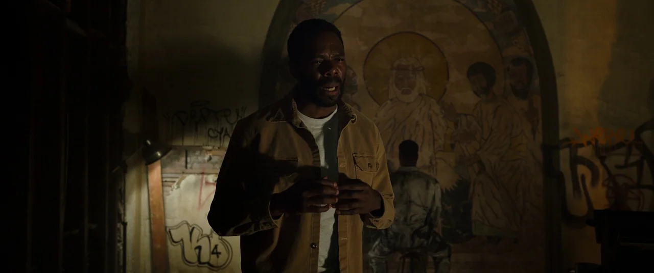 Candyman (2021) movie still: medium — An interior space features crumbling walls covered in spray-painted graffiti and a religious mural. A man in a…