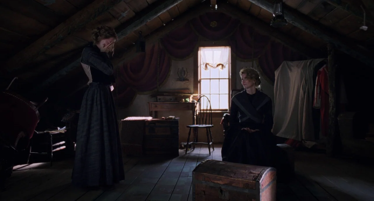 Little Women (2019) movie still: fullbody — A wooden attic space contains a window with heavy drapes and antique furniture. Two women dressed in…