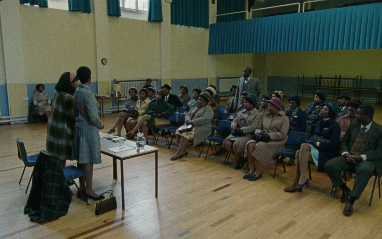 Education (2020) movie still: wide — A spacious community hall with wooden floors and pale yellow walls features a seated audience. Two women stand…