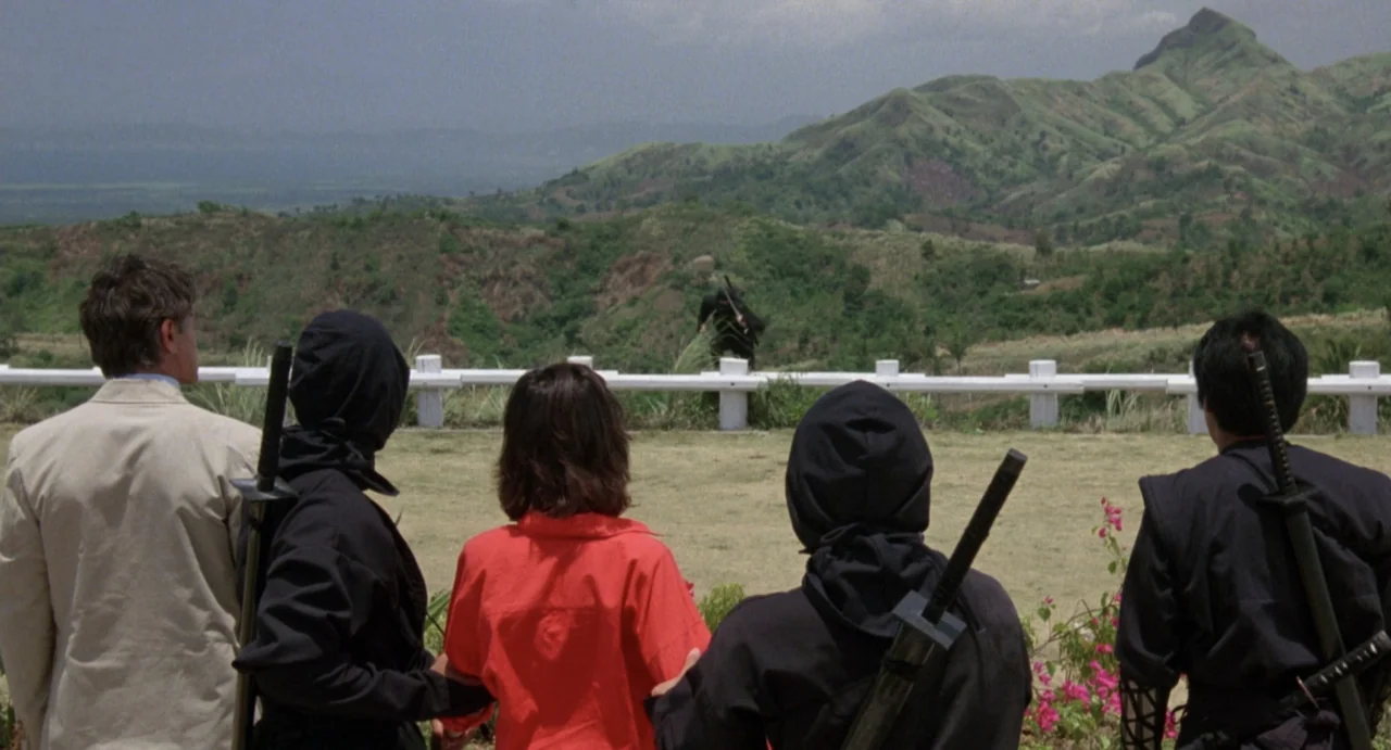 American Ninja (1985) movie still: wide — A grassy lookout point features a white metal railing overlooking distant green hills. A group of five…
