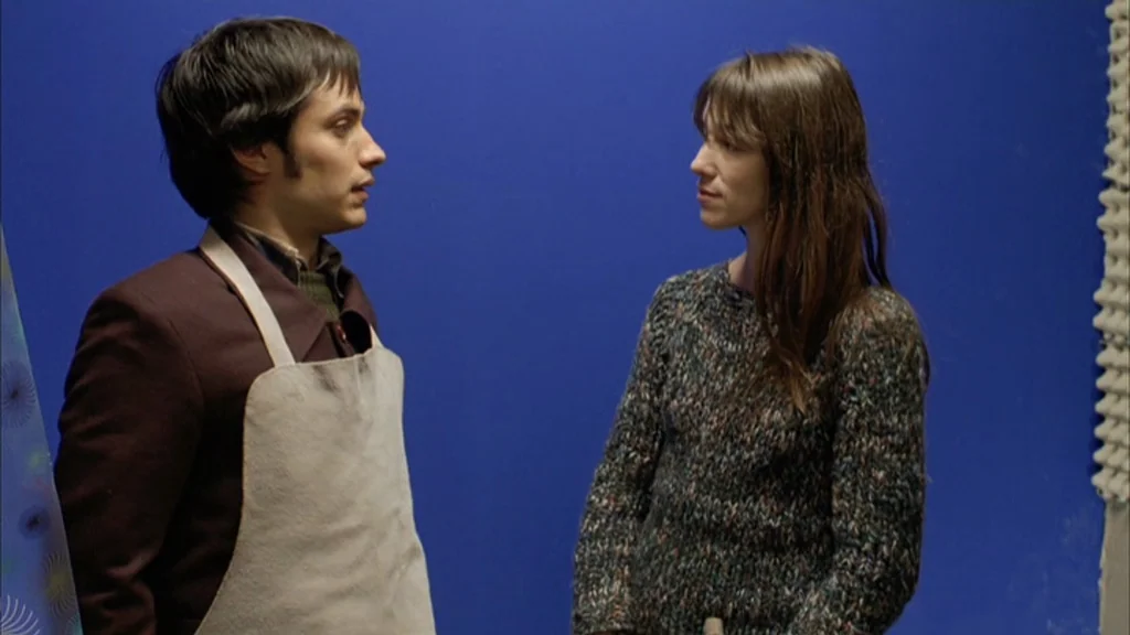 The Science Of Sleep (2006) movie still: medium — Two individuals stand in front of a flat, deep blue wall. A man wearing an apron faces a woman in…