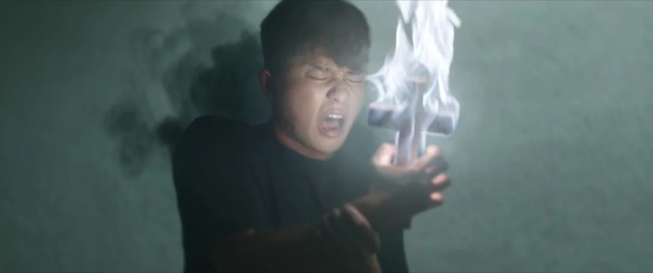 The Divine Fury (2019) movie still: closeup — A dark, featureless room filled with a faint, smoky haze. A young man holds a crucifix that emits an…