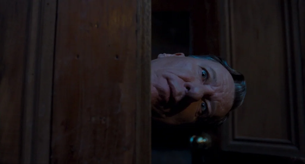 The King’s Speech (2010) movie still: closeup — A man peers through a narrow opening in a dark, wooden door frame. He gazes into the adjacent space…