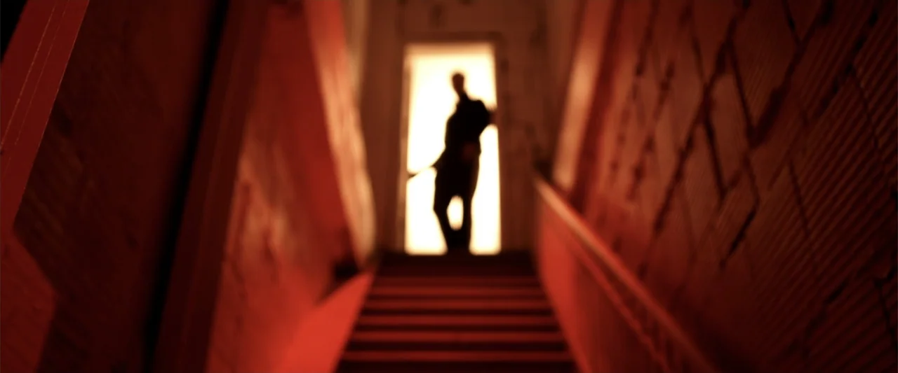 A Walk Among the Tombstones (2014) movie still: wide — A narrow stairwell with red-tinted walls leads upward toward an open, bright doorway. A…