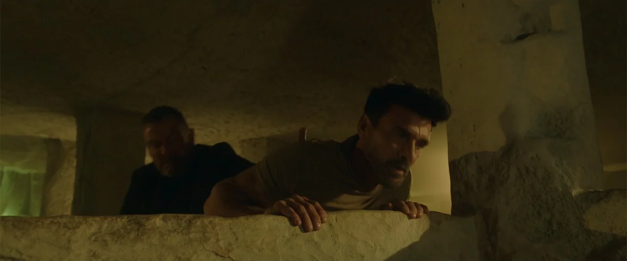 Hounds of War (2024) movie still: medium — A dark, cramped stone tunnel with uneven walls and low ceiling. Two men move cautiously forward through…