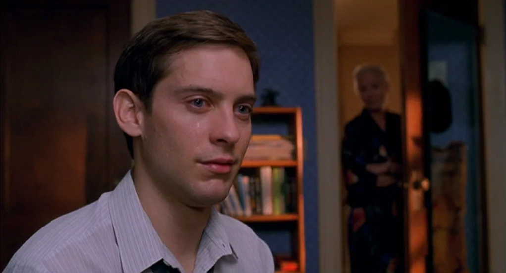 Spider-Man (2002) movie still: closeup — A dimly lit residential interior features blue walls and a wooden bookshelf. A young man sits in the…