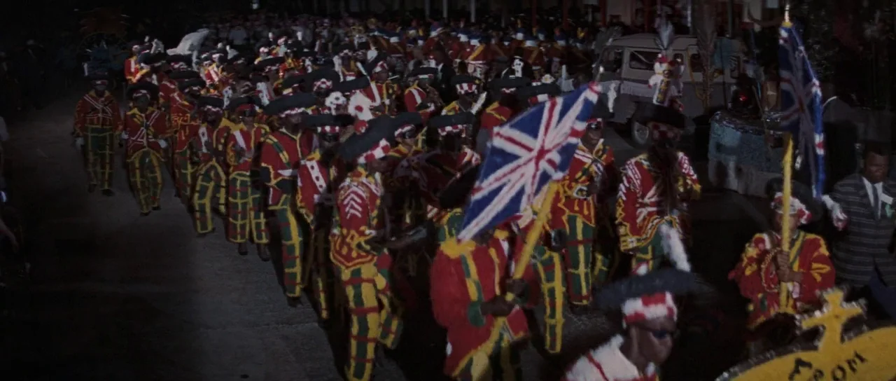Thunderball (1965) movie still: wide — A large group of people marches down a night street during a parade. The participants wear vibrant red,…