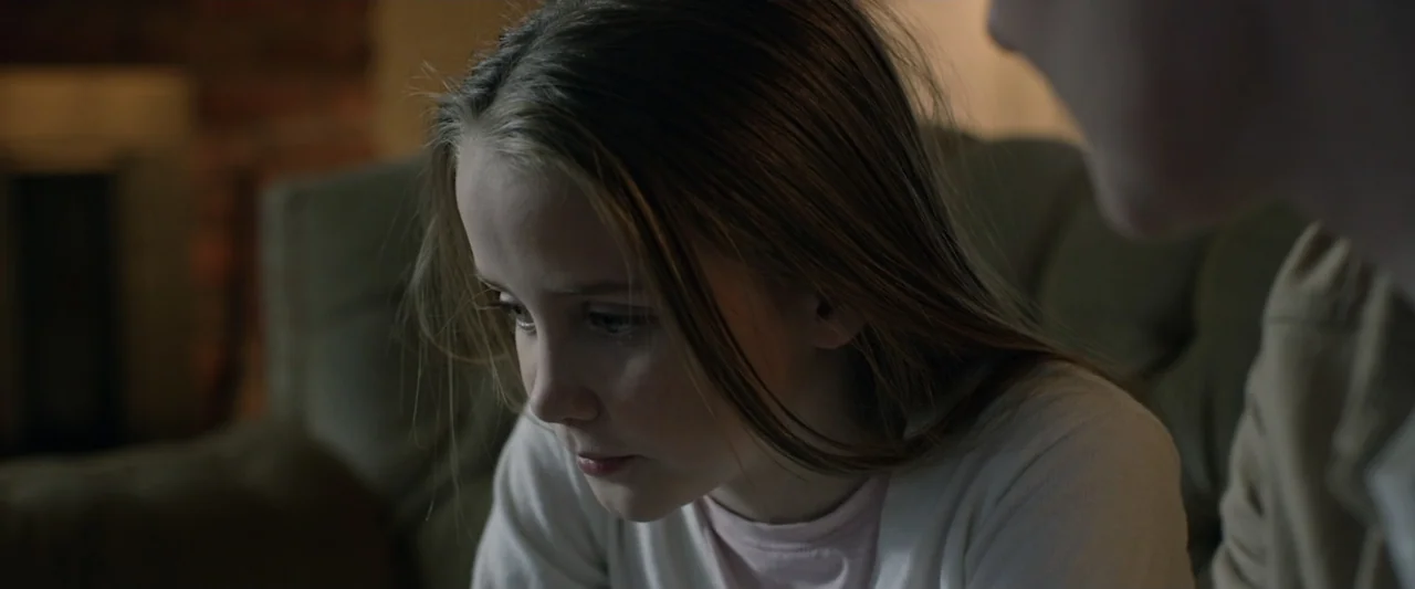 Motherly (2022) movie still: closeup — A domestic space at night with a blurred fireplace visible in the background. A young girl is captured from…